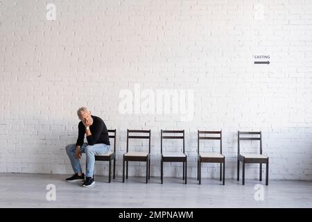 Grauhaariger Schauspieler mittleren Alters, der auf einem Stuhl im Flur sitzt und auf das Casting wartet Stockfoto