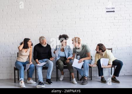 Aufgeregte multirassische Schauspieler mit Szenarien, die sich unterhalten, während sie auf dem Flur sitzen und auf das Casting warten Stockfoto