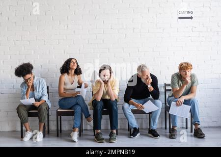 Beunruhigte und durchdachte multiethnische Akteure, die mit Drehbüchern sitzen und auf die Besetzung im Korridor warten Stockfoto