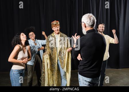 Fröhlicher rothaariger Mann in Königskostüm in der Nähe multiethnischer Schauspieler und Kunstdirektor auf der Bühne im Theater Stockfoto