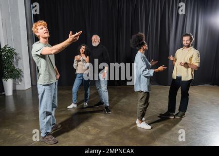 Rothaariger Mann, der mit ausgestreckter Hand neben dem Kunstdirektor probt und multiethnische Schauspieler, die auf der Bühne im Theater sprechen Stockfoto