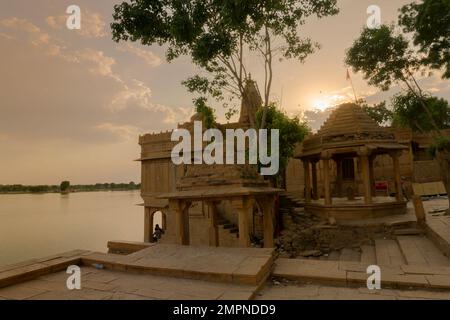 Chhatris und Schreine der hinduistischen Götter und Göttinnen am Gadisar-See, Jaisalmer, Rajasthan, Indien. Indo-islamische Architektur, Sonnenuntergang und bunte Wolken Stockfoto