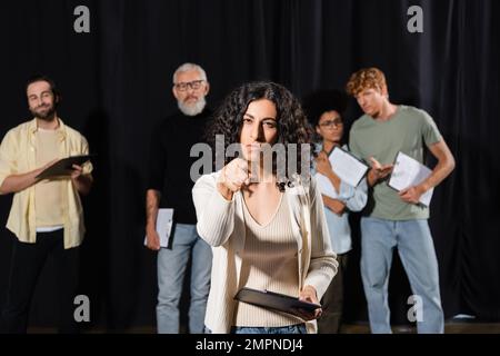 Multirassische Frau mit ernstem Gesichtsausdruck, die auf die Kamera zeigt, während sie in der Nähe von verschwommenen Schauspielern und reifem Kunstdirektor probt Stockfoto