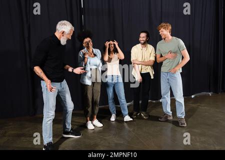 Bärtiger Schauspiellehrer, der mit fröhlichen interrassischen Schülern in der Theaterschule spricht Stockfoto