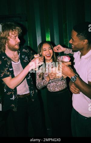 Fröhliche, junge, multiethnische Freunde, die Sandwiches zusammen im Nachtclub essen Stockfoto
