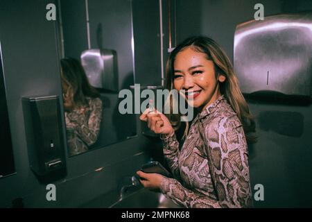 Porträt einer lächelnden jungen Frau, die Lippenstift im Bad im Nachtclub aufträgt Stockfoto