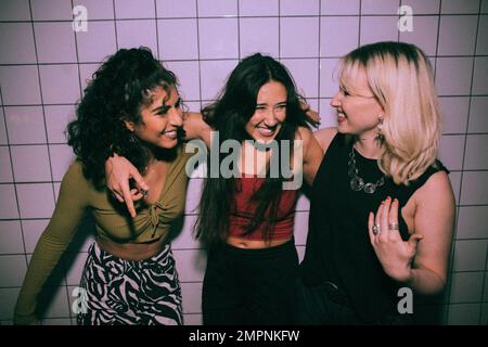 Fröhliche, junge, multirassische, weibliche Freunde, die mit Armen gegen die weiße Wand im Nachtclub stehen Stockfoto