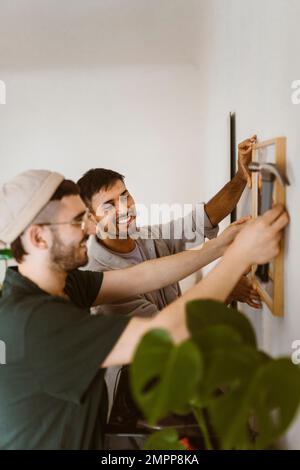Glückliches schwules Paar, das sich gegenseitig hilft, während es zu Hause einen Rahmen an die Wand hängt Stockfoto
