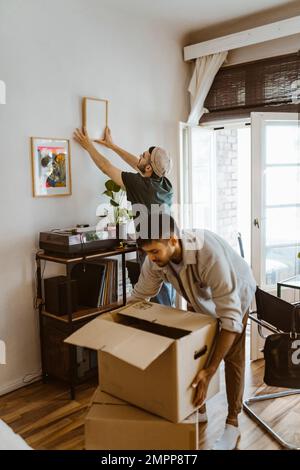 Mann, der Rahmen an der Wand montiert, mit Freund, der zu Hause Kartonboxen stapelt Stockfoto