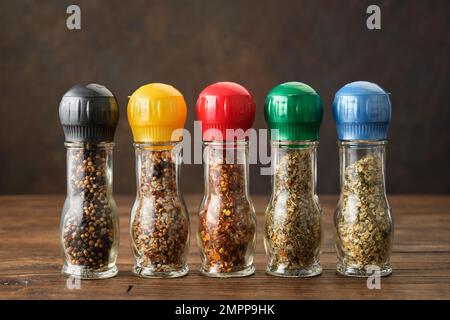 Mühlen mit aromatischen Gewürzen und Kräutern. Mehrere Mahlwerke mit verschiedenen Gewürzen auf Holztisch. Gewürze zum Kochen. Stockfoto