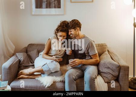 Glückliches Paar, das Kaffee trinkt, während es sein Smartphone mit anderen teilt und zu Hause auf dem Sofa sitzt Stockfoto