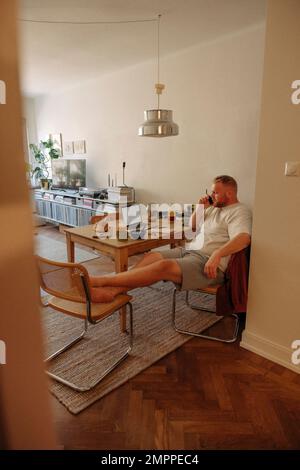 Übergewichtiger Mann, der zu Hause auf einem Stuhl sitzt und mit einem Smartphone spricht Stockfoto