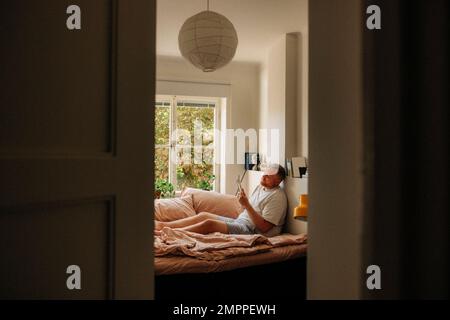 Übergewichtiger Mann liest Buch im Schlafzimmer, durch die Tür gesehen Stockfoto