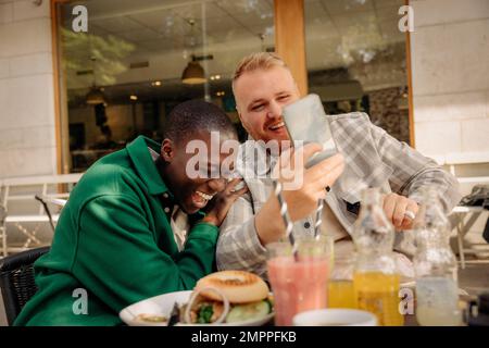 Lächelnder Mann, der sein Smartphone mit einem glücklichen männlichen Freund teilt, während er im Straßencafé sitzt Stockfoto