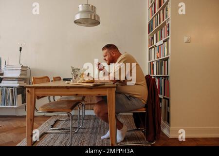 Seitenansicht eines übergewichtigen Mannes, der frühstückt, während er sich den Laptop zuhause ansieht Stockfoto