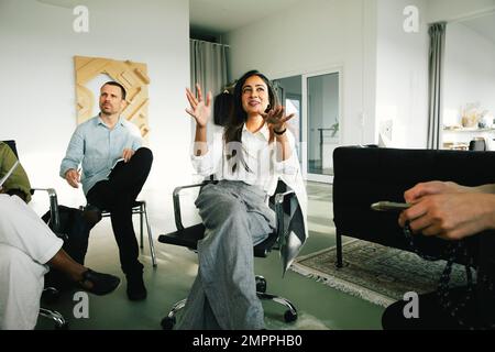 Lächelnde Geschäftsfrau, die Kollegen erklärt, während sie während eines Meetings im Büro auf dem Stuhl sitzt Stockfoto