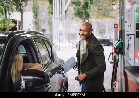 Ein Mann mit rasiertem Kopf, der mit einer Frau spricht, während er an der Tankstelle ein Auto betankt Stockfoto