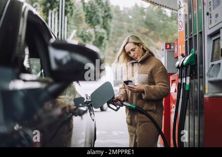 Eine Frau, die ihr Handy benutzt, während sie an der Tankstelle ihr Auto betankt Stockfoto