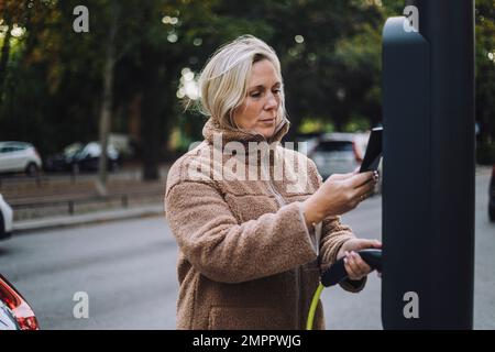 Frau, die das Ladegerät hält, während sie das Smartphone an der Ladestation durchsucht Stockfoto