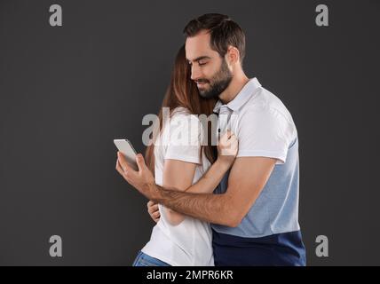 Ein Mann, der sich für ein Smartphone interessiert, während er seine Freundin auf schwarzem Hintergrund umarmt. Beziehungsprobleme Stockfoto