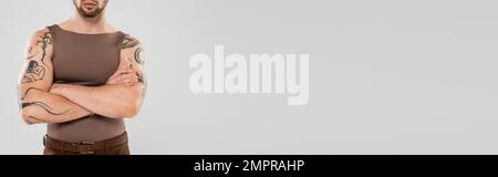 Zuschnittsansicht eines jungen tätowierten Mannes, der Arme überkreuzt, isoliert auf grauem Banner Stockfoto
