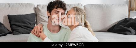 Glückliche blonde Frau küsst die Wange des Freundes im Wohnzimmer, Banner, Stock Image Stockfoto
