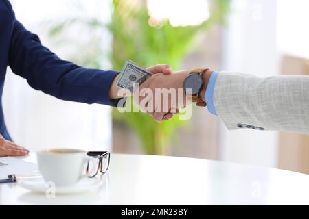 Mann und Frau, die sich die Hände mit Dollar zwischen den Handflächen schütteln, an Tisch drinnen, Nahaufnahme. Bestechungskonzept Stockfoto