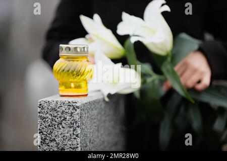 Eine Frau, die weiße Lilien in der Nähe von grauen Granitgräbern hält, konzentriert euch auf Kerzen. Beerdigungszeremonie Stockfoto