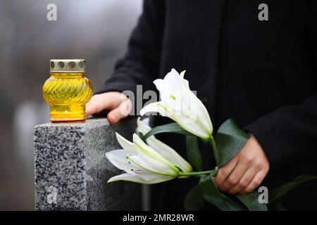 Eine Frau, die weiße Lilien in der Nähe von grauen Granitgräbern hält, mit Kerzen im Freien, Nahaufnahme. Beerdigungszeremonie Stockfoto