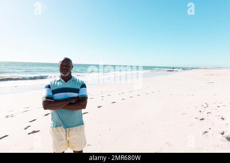 Ein afroamerikanischer, glatzköpfiger Senior-Mann mit gekreuzten Armen, der am Sandstrand vor klarem Himmel stand Stockfoto