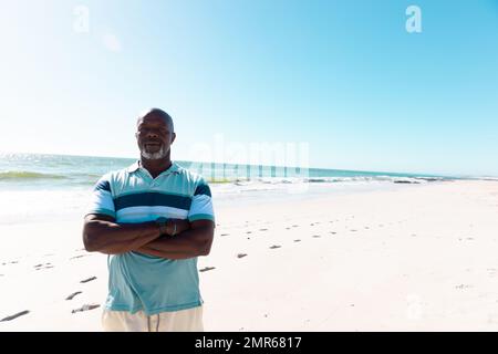 Kahlköpfiger afroamerikanischer Seniorenmann mit gekreuzten Armen, der am Strand gegen das ruhige Meer und den Himmel steht Stockfoto