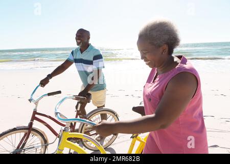 Lächelndes afroamerikanisches Seniorenpaar mit Fahrrädern, das im Sommer am Strand gegen Meer und Himmel spaziert Stockfoto