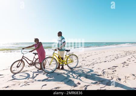 afroamerikanisches Seniorenpaar mit Fahrrädern, die im Sommer am Strand vor klarem Himmel spazieren und sich unterhalten Stockfoto