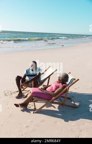 afroamerikanisches Seniorenpaar, das auf Liegestühlen sitzt und sich im Sommer am Strand unter klarem Himmel unterhält Stockfoto