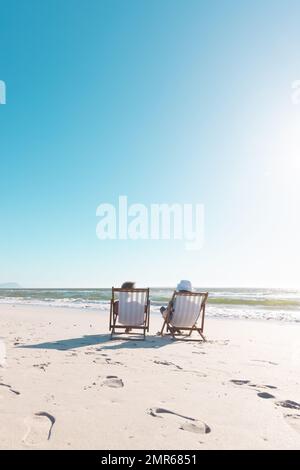 afroamerikanisches Seniorenpaar, das sich auf Liegestühlen vor dem Meer unter klarem blauen Himmel entspannt Stockfoto