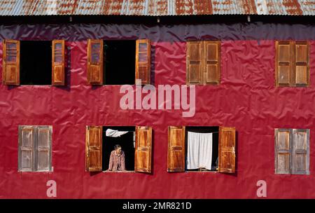 Eine Frau blickte aus dem Fenster eines alten roten Hauses mit abgenutzten Holzläden in Myanmar (Birma). Stockfoto