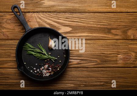 Schwarzer Bratpfanne aus Gusseisen mit Speisezutaten frischer Rosmarin, Knoblauch, Salz und Pfeffer auf rustikalem Holztisch, Kochhintergrund und Stockfoto