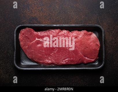 Rind mageres Filet Steak in schwarzem Kunststoffbehälter auf dunkelbraunem rustikalem Betonhintergrund von oben flach liegend, Diät Rindfleisch Steak fertig für C Stockfoto