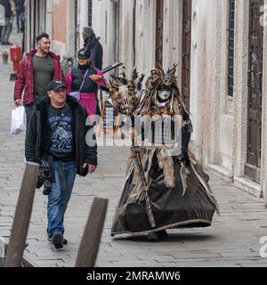 Ein Fotograf geht mit einem Modell in Karnevalskostüm einer heidnischen Hexe an der Straße vorbei, mit Möbeln, Pelzen und Federn darauf in Venedig, Italien Stockfoto