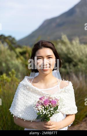 Vertikales Porträt einer glücklichen asiatischen Braut, die einen Strauß hält, der bei einer Hochzeit im Freien lächelt, Kopierbereich Stockfoto