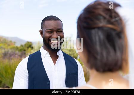 Frohes afroamerikanisches Bräutigam, das die vielseitige Braut anlächelt, bei einer Hochzeit im Freien, Kopierbereich Stockfoto