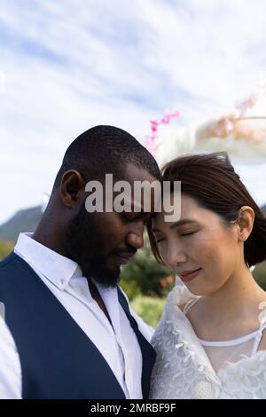 Vertikale von glücklichen, vielfältigen Paaren, die sich mit Köpfen berühren, mit geschlossenen Augen bei einer Hochzeit im Freien, Kopierbereich Stockfoto