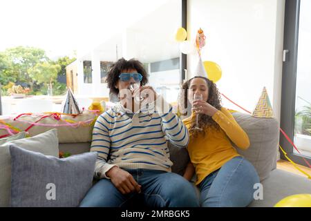 Glückliches birassisches Paar, das zu Hause Party macht, auf der Couch sitzt und Champagner trinkt, Kopierraum Stockfoto