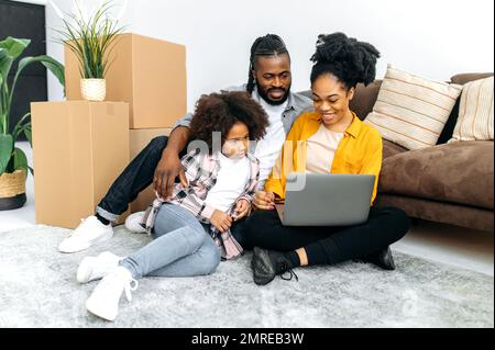 Glückliches afroamerikanisches Ehepaar mit Vorschultochter, sitzen neben einem Sofa im Wohnzimmer zwischen Pappkartons in ihrem neuen Zuhause, verwenden Laptop, verbringen Zeit zusammen beim Online-Shopping, lächeln Stockfoto