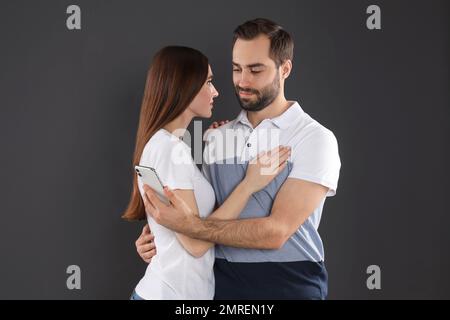 Ein Mann, der sich für ein Smartphone interessiert, während er seine Freundin auf schwarzem Hintergrund umarmt. Beziehungsprobleme Stockfoto