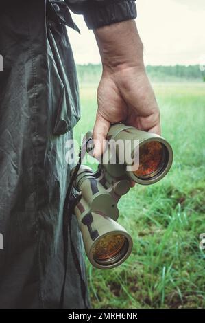 Nahaufnahme der Hand eines Jägers oder Reisenden oder Grenzschutzbeamten in dunklem Khaki-Regenmantel mit grünem Fernglas und roter Linse vor dem Hintergrund Stockfoto