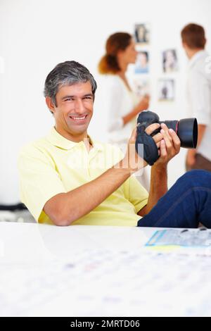 Fotograf checkt Bilder in der Kamera aus. Porträt eines glücklichen Fotografen, der sich Bilder in seiner Kamera mit Kollegen im Hintergrund ansieht. Stockfoto