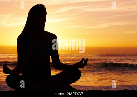 Silhouette Frau in Lotusposition gegen das Meer bei Sonnenuntergang. Silhouettenbild einer Frau, die bei Sonnenuntergang in Lotusposition gegen das Meer meditiert. Stockfoto