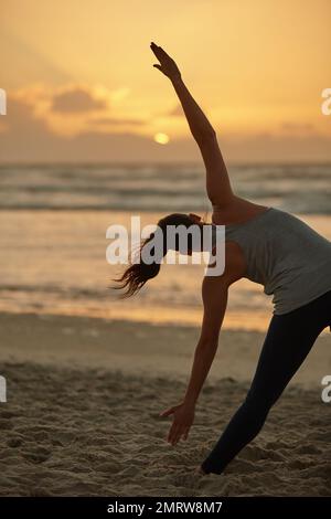 Ihre tägliche körperliche Routine in der Natur. Rückansicht einer Frau, die bei Sonnenuntergang am Strand Yoga macht. Stockfoto