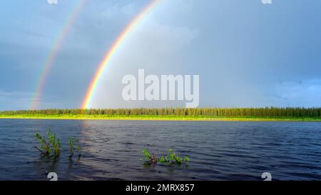Teil eines doppelten Regenbogens vor dem Hintergrund von hellen Wolken und einem sonnenbeleuchteten Wald auf der anderen Seite des Flusses Vilyuy mit Regentropfen im Wasser Stockfoto